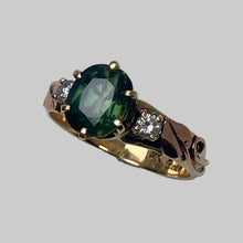 Load image into Gallery viewer, A three-stone ring featuring a large oval-cut natural dark green Sapphire in the center, flanked by two round clear recycled diamonds, set on a yellow gold band with a pair of rose gold leaf and vines.
