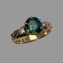 Load image into Gallery viewer, A three-stone ring featuring a large oval green Sapphire in the center, flanked by two small round diamonds, set on a yellow gold band with 2 rose gold leaves and vines.
