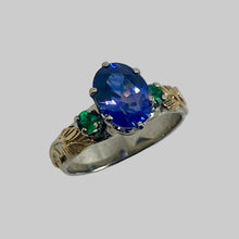 Load image into Gallery viewer, A vintage-style ring featuring a large oval-cut purple/blue sapphire center stone, accented by a ound emerald either side, set in a 14K white gold band with 18L yellow gold leaf and vine details.
