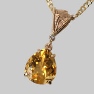A large citrine pendant with a pear shape, set in a gold-colored frame with a bezel-set diamond in the background, suspended by a gold chain.